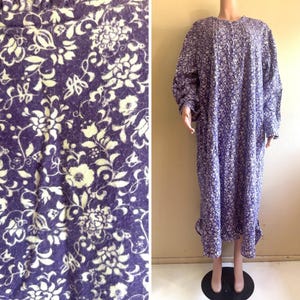 May include: A long-sleeved, floor-length dress in a deep purple hue featuring a dense, white floral pattern throughout. The garment includes a round neckline with a button-down placket, long sleeves with gathered cuffs, and a ruffled hemline detail.