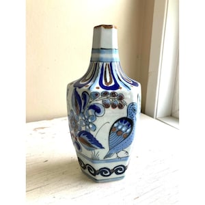May include: A hexagonal ceramic vase featuring a white base with hand-painted blue and brown floral patterns and a bird motif. The neck is narrow and cylindrical, transitioning into a wider, six-sided body decorated with scrolling blue lines.