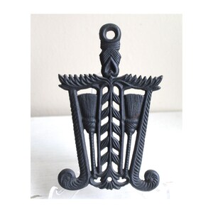 Op de afbeelding: Een zwarte gietijzeren trivet met een ontwerp van drie bezems en een bladmotief. De trivet heeft een decoratieve bovenkant met een hartvorm.
