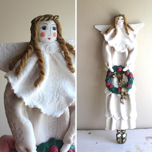 Puede incluir: Una figura de ángel de cerámica blanca con alas detalladas y un vestido fluido. El ángel tiene cabello largo y castaño trenzado y sostiene una corona verde adornada con rosas rojas y bayas. La figura tiene un halo dorado.