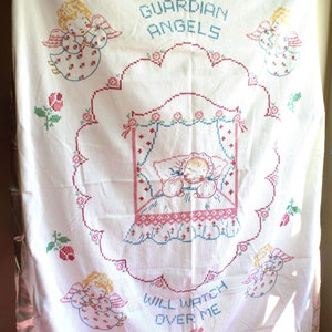 May include: White embroidered baby blanket with pink satin trim. The blanket features a central design of a sleeping baby in a crib with angels surrounding the crib. The text "Guardian Angels Will Watch Over Me" is embroidered on the blanket.