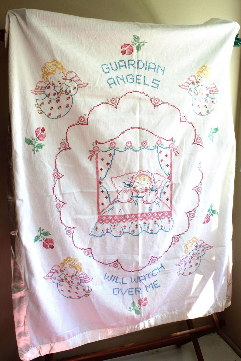 May include: White embroidered baby blanket with pink satin trim. The blanket features a central design of a sleeping baby in a crib with angels surrounding the crib. The text "Guardian Angels Will Watch Over Me" is embroidered on the blanket.