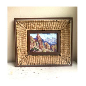 May include: A framed landscape painting of a mountain range. The painting is set within a dark brown wooden frame, which is surrounded by a woven, light brown, rectangular frame. The painting depicts red and brown rock formations, green trees, and a blue sky.