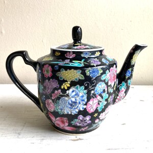 Może przedstawiać: Czarny ceramiczny czajnik z pokrywką, uchwytem i dzióbkiem. Czajnik zdobiony jest kolorowym wzorem kwiatowym z różowymi, niebieskimi, żółtymi i fioletowymi kwiatami. Uchwyt i dzióbek są czarne, a pokrywka ma czarny guz.