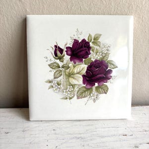 Op de afbeelding: Een vierkante witte keramische tegel met een centrale bloemenillustratie van twee dieppaarse rozen, een rozenknop, groene bladeren en kleine witte vulbloemen. Het ontwerp is gedrukt op een glad, glanzend oppervlak met strakke randen.