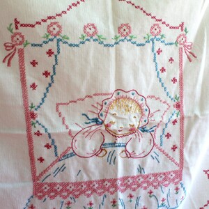 May include: White cotton fabric with pink, blue, and green embroidery of a sleeping baby in a crib. The embroidery is done in a cross-stitch pattern.