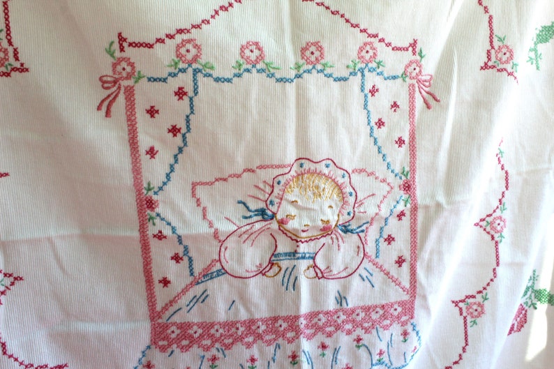 May include: White cotton fabric with pink, blue, and green embroidery of a sleeping baby in a crib. The embroidery is done in a cross-stitch pattern.