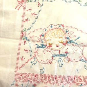 May include: A white embroidered baby blanket with a pink and blue floral design. The center of the blanket features a hand-stitched image of a baby girl with a pink bonnet and blue dress. The blanket is edged with a pink and blue stitched border.
