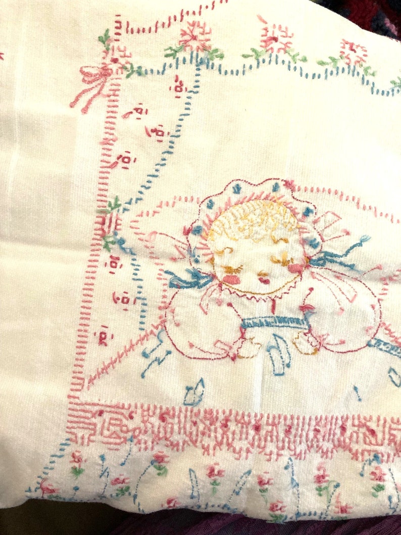 May include: A white embroidered baby blanket with a pink and blue floral design. The center of the blanket features a hand-stitched image of a baby girl with a pink bonnet and blue dress. The blanket is edged with a pink and blue stitched border.