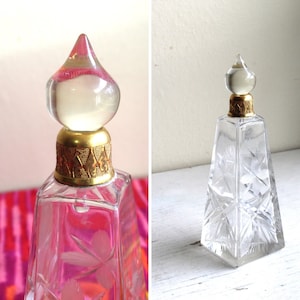 Puede incluir: Un frasco de perfume de vidrio transparente con un tapón dorado y un diseño intrincado de vidrio cortado. El frasco tiene forma de pirámide y está lleno de un líquido transparente.