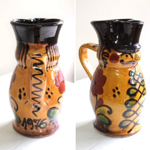 Peut inclure: Un pichet en céramique vintage avec un bord et une anse noirs, et un corps jaune décoré de fleurs rouges, de feuilles vertes et d'accents noirs. L'année "1976" est peinte sur le devant. Le pichet a un aspect unique et artisanal.