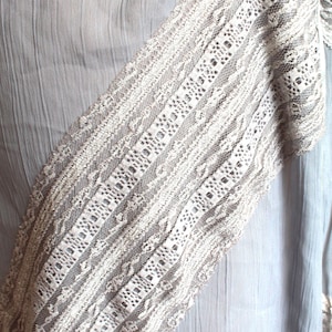 May include: A light grey fabric with a white lace trim. The lace has a delicate pattern and is sewn onto the fabric in a diagonal line.