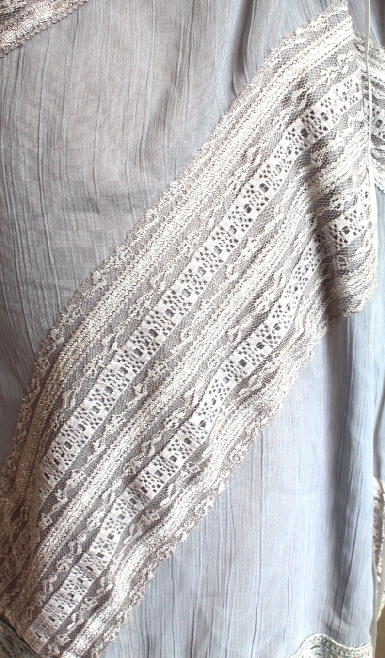 May include: A light grey fabric with a white lace trim. The lace has a delicate pattern and is sewn onto the fabric in a diagonal line.