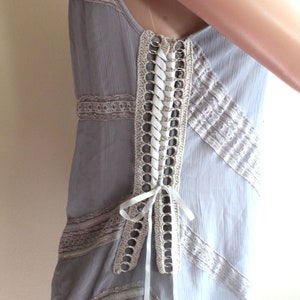 May include: A grey sleeveless dress with a white lace overlay and a white ribbon tied at the back. The dress has a crochet detail with metal rings along the back.