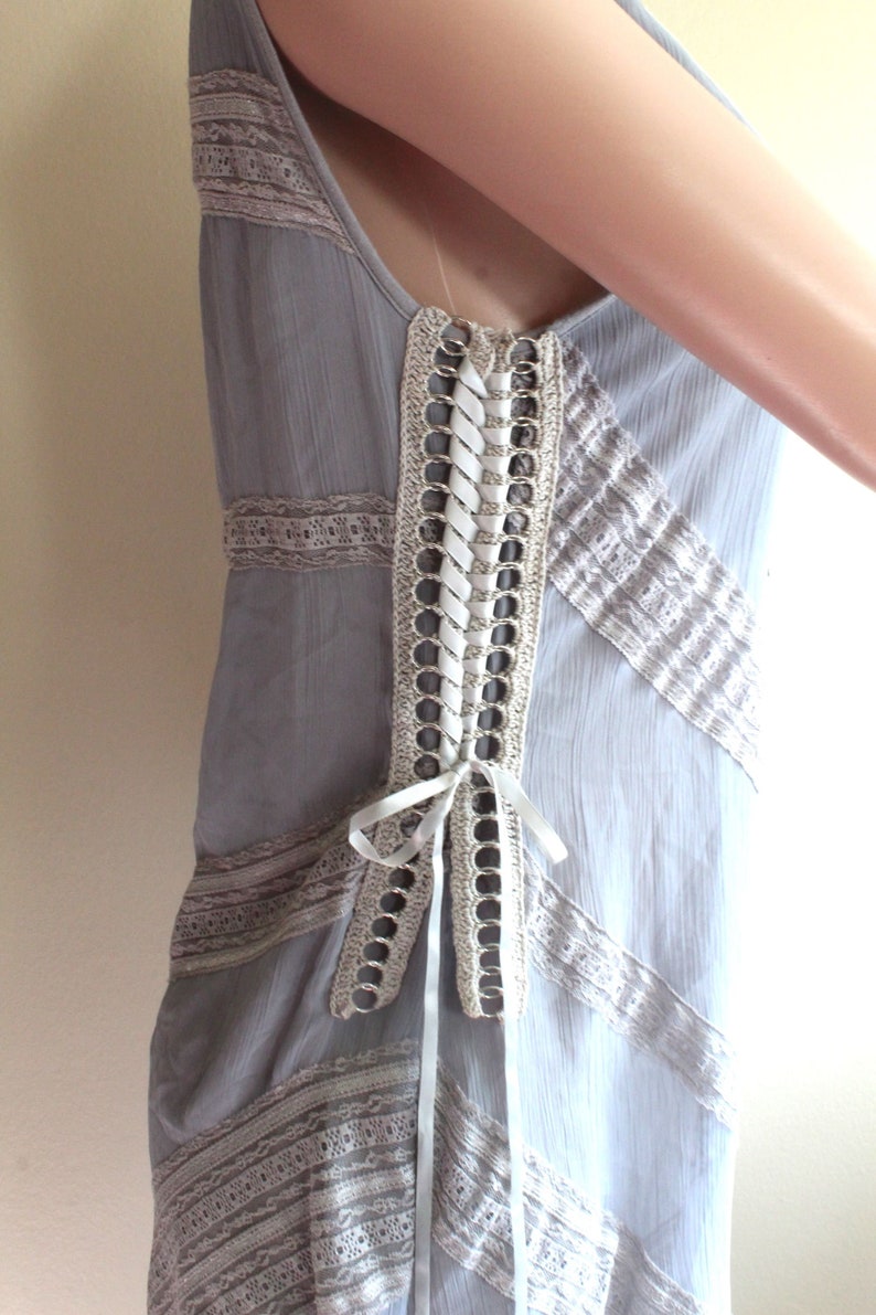 May include: A grey sleeveless dress with a white lace overlay and a white ribbon tied at the back. The dress has a crochet detail with metal rings along the back.