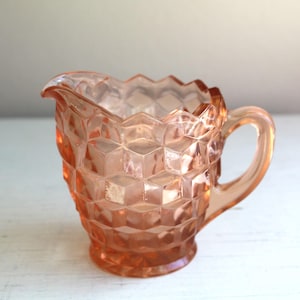 Peut inclure: Un pot à crème en verre rose avec un motif en diamant. Le pot à crème a une poignée et une base arrondie.