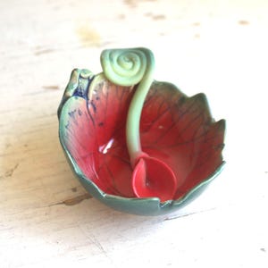 May include: A ceramic leaf-shaped dish with a red interior and a green rim. A green, spiral-shaped handle extends from the dish, with a red spoon resting inside. The dish is on a white surface.