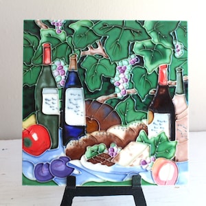 May include: A ceramic tile featuring a painted scene of wine bottles, grapes, bread, and cheese. The tile is green, blue, red, and brown. It is displayed on a black easel.