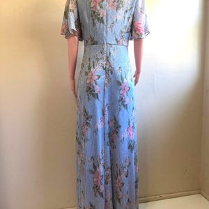 Op de afbeelding: Lichtblauwe, vloerlange jurk met een bloemenpatroon. De jurk heeft korte, fladderende mouwen en een V-hals. Roze en witte bloemen zijn gedrukt op de jurk, die een verticaal gestreepte textuur heeft.