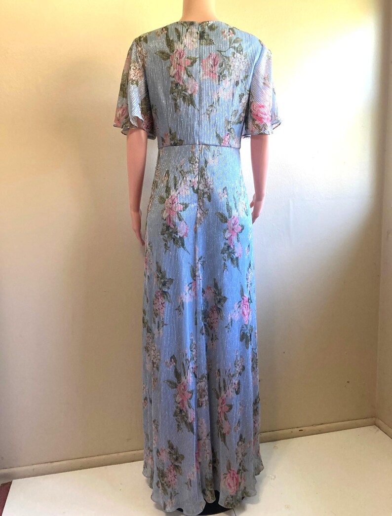 Op de afbeelding: Lichtblauwe, vloerlange jurk met een bloemenpatroon. De jurk heeft korte, fladderende mouwen en een V-hals. Roze en witte bloemen zijn gedrukt op de jurk, die een verticaal gestreepte textuur heeft.