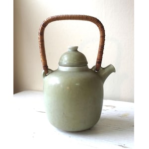 May include: A sage green ceramic teapot with a woven rattan handle. The teapot has a rounded body, a small spout, and a matching lid. The handle arches over the top, connecting to the body on either side. The teapot is likely vintage or handmade.