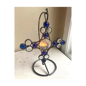 Op de afbeelding: Een decoratieve metalen kruisvormige kandelaar. Het kruis heeft blauwe en heldere glazen accenten en bevat een brandende waxinelichtkaars. De houder heeft een cirkelvormige basis en een gebogen bovenkant om op te hangen.