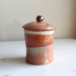 May include: A ceramic jar with a lid, featuring a brown, white and orange glaze. The jar has a rounded shape and a smooth, slightly textured surface.
