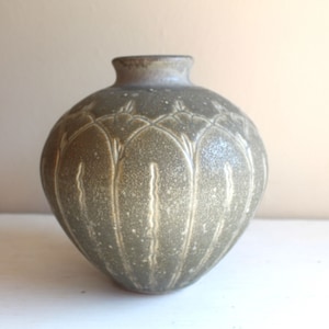 May include: A small, round, green ceramic vase with a textured surface and a decorative design. The vase has a slightly flared lip and a rounded base.
