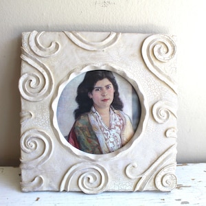 May include: Square, off-white picture frame with a portrait of a woman. The frame has a textured, crackled finish and ornate, swirling scrollwork. The portrait features a woman with dark hair and a colorful shawl.