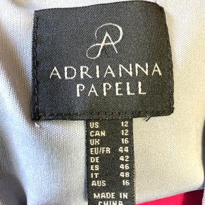 Op de afbeelding: Een zwart label met de merknaam "ADRIANNA PAPELL" in witte tekst. Het label bevat maatconversies voor US, CAN, UK, EU/FR, DE, ES, IT en AUS. Het label leest "MADE IN CHINA".