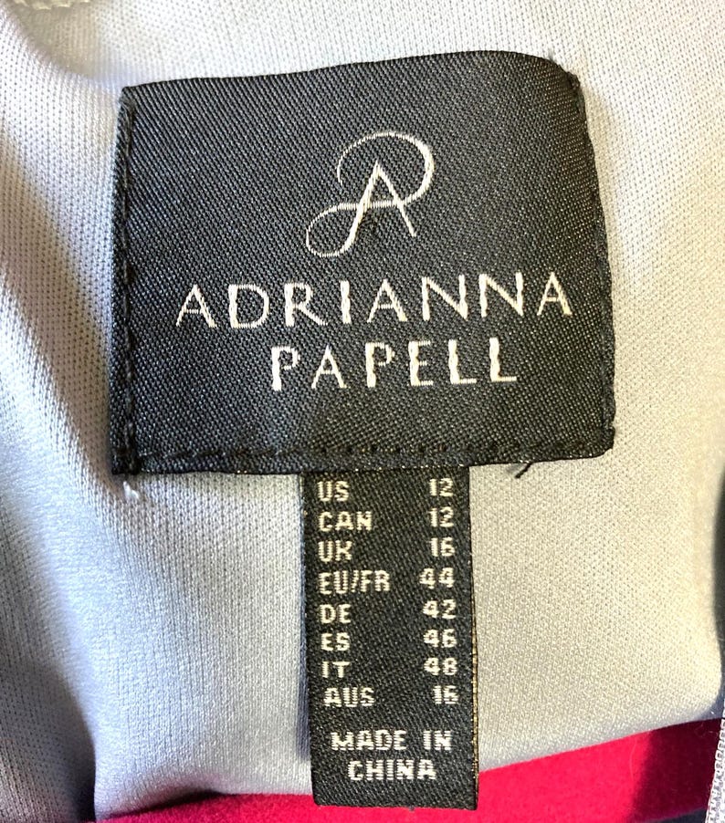 Op de afbeelding: Een zwart label met de merknaam "ADRIANNA PAPELL" in witte tekst. Het label bevat maatconversies voor US, CAN, UK, EU/FR, DE, ES, IT en AUS. Het label leest "MADE IN CHINA".