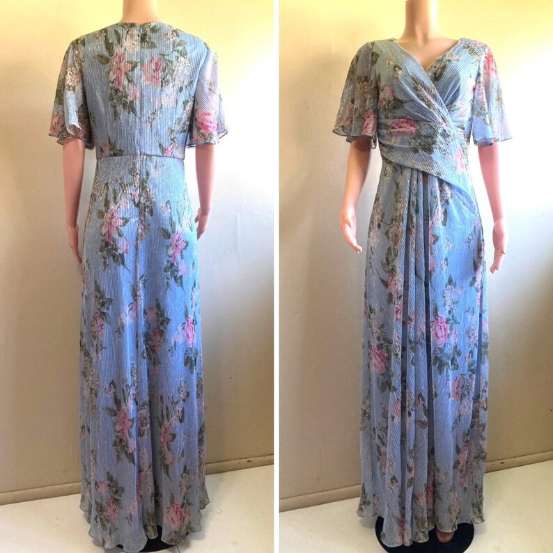 Op de afbeelding: Een lichtblauwe, vloerlange jurk met een bloemenpatroon van roze en witte bloemen. De jurk heeft korte, fladderende mouwen en een V-hals. De stof heeft verticale strepen en de jurk wordt op een mannequin getoond.