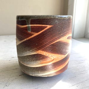 May include: A cylindrical ceramic cup featuring a textured, dark brown and reddish-orange swirled pattern across its exterior. The rim has a smooth, greyish-brown glaze with a single drip mark running down the side, creating a distinct contrast.