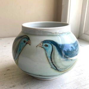 Puede incluir: Un pequeño jarrón de cerámica redondeado con base blanca y acabado liso y brillante. El exterior presenta ilustraciones pintadas a mano de dos pájaros azules con contornos oscuros, colocados uno frente al otro. Tiene un borde circular ancho.