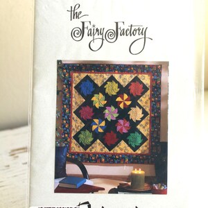 Puede incluir: Un paquete con un patrón de edredón con diseños de molinillos. El edredón tiene un fondo negro con molinillos vibrantes en varios colores. El texto "The Fairy Factory" y "Origami" son visibles.
