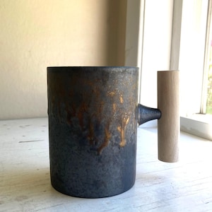 May include: A cylindrical mug with a dark, mottled charcoal and black finish featuring metallic gold-toned drips running down the exterior surface. The mug has a straight, light-colored wooden handle attached to the side with a dark metal connector.