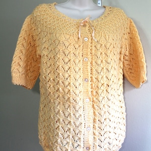 Puede incluir: Un cárdigan de crochet amarillo con cierre de botones y mangas cortas. El cárdigan tiene un delicado patrón de encaje y un lazo en el escote.