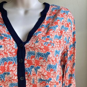 May include: Coral-colored button-down shirt with a blue tiger print and white floral accents. The shirt has a navy blue trim along the V-neck and button placket. The long-sleeved shirt is displayed on a mannequin.