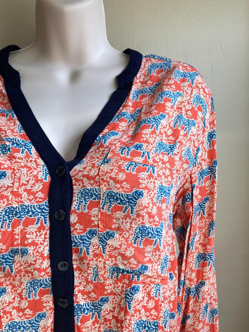 May include: Coral-colored button-down shirt with a blue tiger print and white floral accents. The shirt has a navy blue trim along the V-neck and button placket. The long-sleeved shirt is displayed on a mannequin.