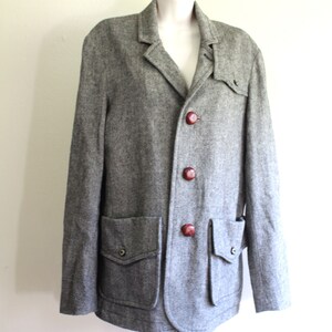 May include: A grey tweed jacket with a button fastening and two large pockets. The jacket has brown buttons and a slightly rounded hem.