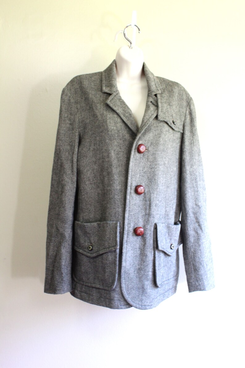 May include: A grey tweed jacket with a button fastening and two large pockets. The jacket has brown buttons and a slightly rounded hem.