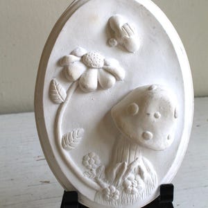 May include: White ceramic oval wall plaque with a raised design of a mushroom, a flower, and a butterfly. The plaque is on a black easel.