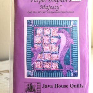 Op de afbeelding: Een quiltpatroon getiteld "Purple Dolphin's Majesty" in een plastic hoes. Het patroon toont een paars dolfijnontwerp op een blauwe en paarse achtergrond. De quiltgrootte is 152 cm x 168 cm. Het pakket bevat ook het Java House Quilts-logo.