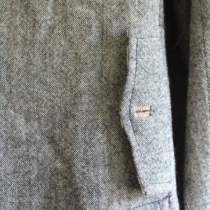 May include: A close-up of a grey tweed jacket with a small pocket on the left side. The pocket has a brown button.
