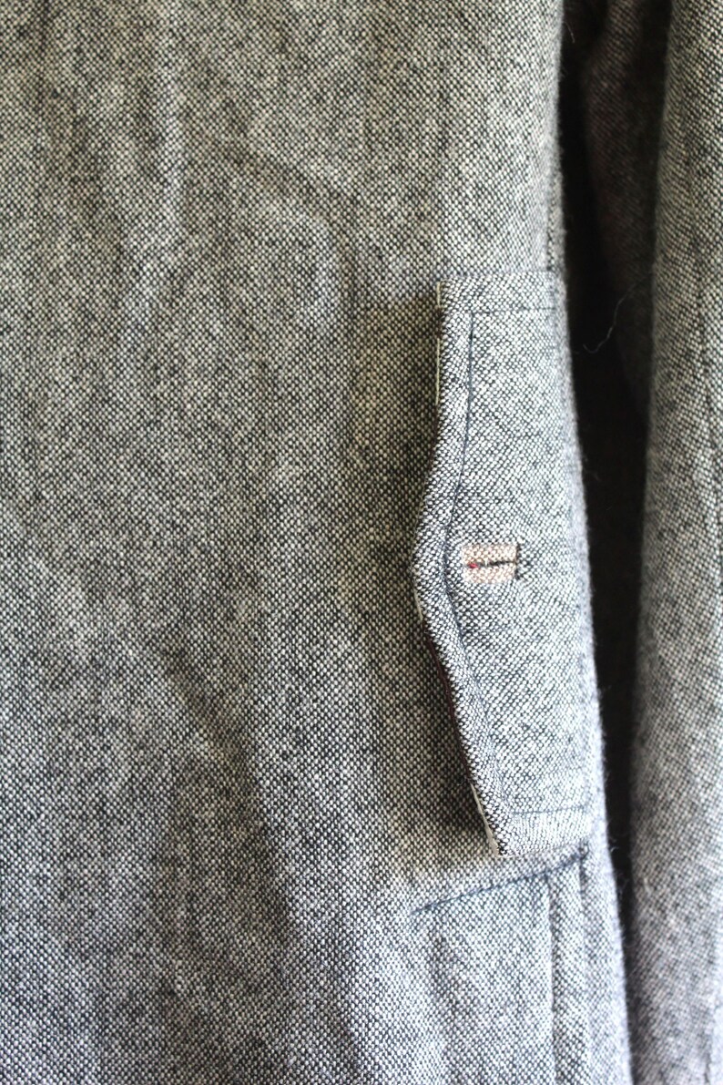 May include: A close-up of a grey tweed jacket with a small pocket on the left side. The pocket has a brown button.