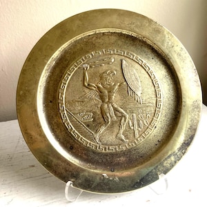 May include: A round, antique brass plate with a detailed relief of a warrior figure. The warrior is depicted in action, holding a weapon and shield, surrounded by a Greek key border and a desert landscape. The plate has a worn, aged appearance.
