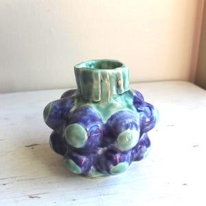 Könnte beinhalten: Eine handgefertigte Keramikvase mit einem einzigartigen, strukturierten Design. Die Vase hat einen bauchigen Körper mit abgerundeten Vorsprüngen in Blau- und Grüntönen. Die Öffnung ist in einem helleren Grünton gehalten. Die Vase ist ein dekorativer Gegenstand.