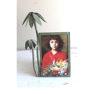 May include: A vintage metal palm tree-shaped picture frame. It holds a portrait of a young girl in a red dress holding flowers. The frame has a distressed, aged appearance. The portrait is rectangular.