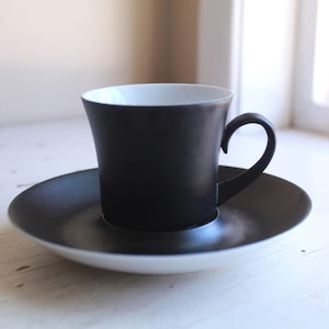 May include: A black coffee cup with a matching saucer. The cup has a curved handle and a white interior. The saucer is round and the same black color as the cup. The set is placed on a white surface.