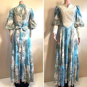 May include: A vintage floor-length dress with a blue and white toile pattern. The dress features long, puffy sleeves, a high neckline, and a cream-colored lace bodice. A matching sash ties at the waist. The dress is displayed on a mannequin.